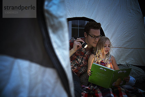 Vater hält Taschenlampe und liest der Tochter vor  während er im Zelt sitzt