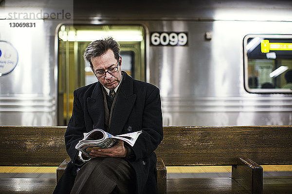 Geschäftsmann liest Zeitschrift  während er auf einer Bank am Bahnhof sitzt