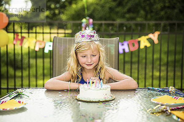 Lächelndes Mädchen mit Krone schaut auf Geburtstagskuchen auf dem Tisch im Hinterhof