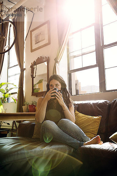 Frau in voller Länge  die an einem sonnigen Tag Tee trinkt  während sie sich zu Hause auf dem Sofa ausruht