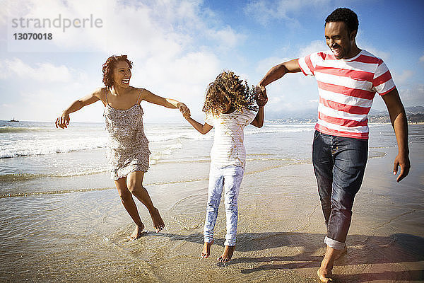 Verspielte Familie hält die Hände der Tochter  während sie sich am Strand am Ufer vergnügt
