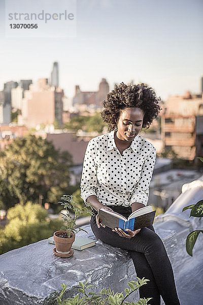 Lächelnde Frau liest Buch  während sie auf einer Gebäudeterrasse gegen den Himmel sitzt