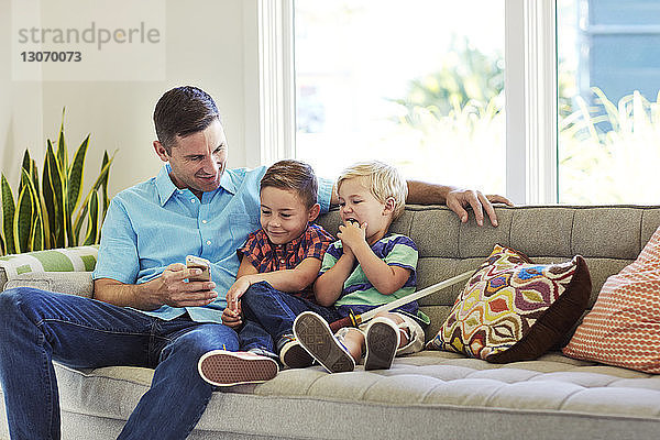 Mann sitzt mit Söhnen auf dem Sofa und schaut auf Handy