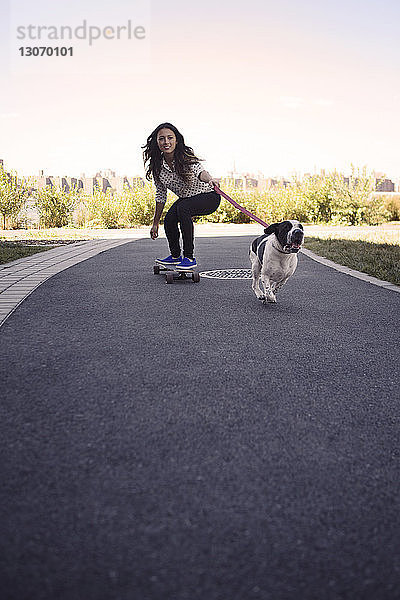Frau fährt Skateboard mit laufendem Hund auf der Straße gegen den klaren Himmel