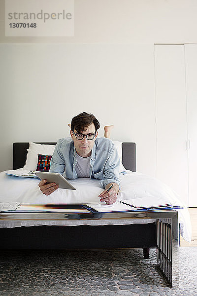 Portrait eines Mannes  der einen Tablet-Computer benutzt  während er zu Hause am Bett an der Wand arbeitet