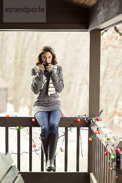 Porträt einer Frau  die einen Drink genießt  während sie auf einem Geländer in der Veranda sitzt