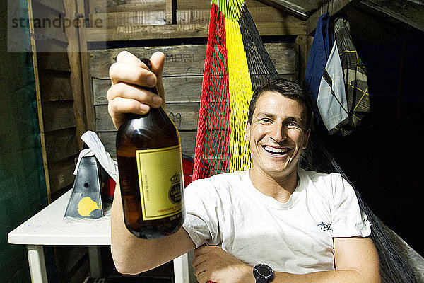 Porträt eines glücklichen Mannes  der eine Bierflasche in der Hand hält  während er sich auf einer Hängematte in einer Blockhütte ausruht