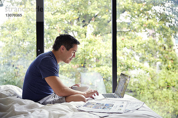 Seitenansicht eines Mannes mit Laptop beim Frühstück am Bett am Fenster