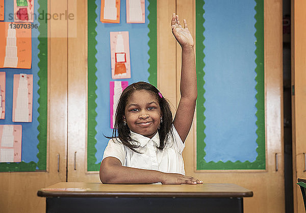 Porträt eines Mädchens  das die Hand hebt  während es am Schreibtisch im Klassenzimmer sitzt