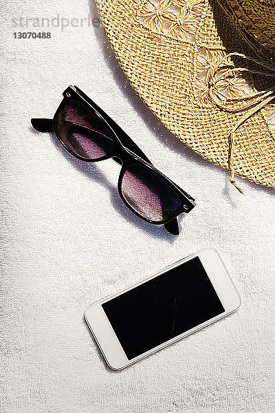 Draufsicht auf Sonnenbrille und Smartphone mit Strohhut auf einem Handtuch am Strand