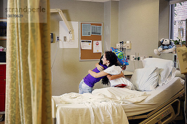 Mutter umarmt Mädchen  während sie im Krankenhaus auf dem Bett sitzt