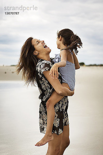 Glückliche Mutter trägt Tochter  während sie am Strand im Sand steht