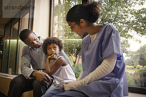 Arzt spricht mit Mädchen  das mit seinem Vater auf dem Fensterbrett im Krankenhaus sitzt