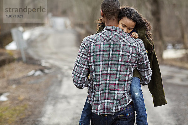 Rückansicht eines Vaters  der seine Tochter trägt  während er auf einer Straße im Wald geht
