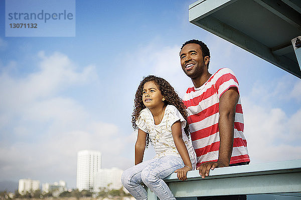 Vater und Tochter schauen weg  während sie sich am Geländer am Pier gegen den Himmel entspannen