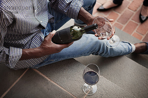 Hochwinkelaufnahme eines Mannes  der Rotwein ausschenkt  während er auf einer Treppe sitzt