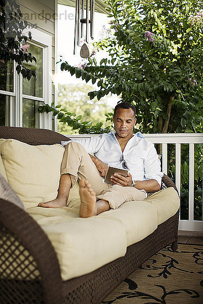 Mann benutzt Tablet-Computer  während er sich auf dem Sofa in der Veranda entspannt