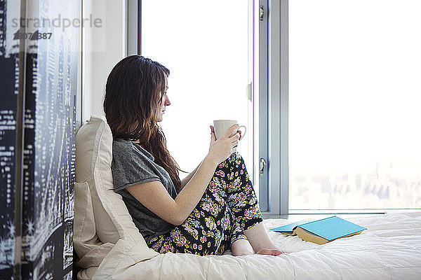 Seitenansicht einer Frau mit Kaffeetasse  die wegschaut  während sie zu Hause auf dem Bett sitzt