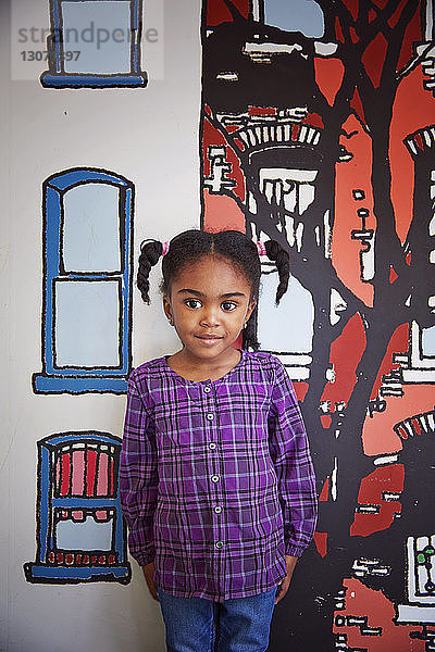 Porträt eines niedlichen Mädchens  das an einer bemalten Wand im Kindergarten steht
