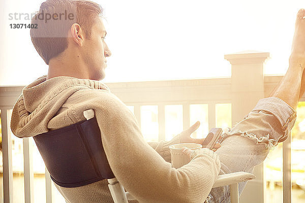 Nachdenklicher Mann hält Smartphone in der Hand  während er auf dem Balkon sitzt