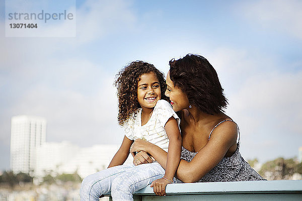 Frau umarmt Tochter  während sie sich am Geländer am Pier gegen den Himmel entspannt