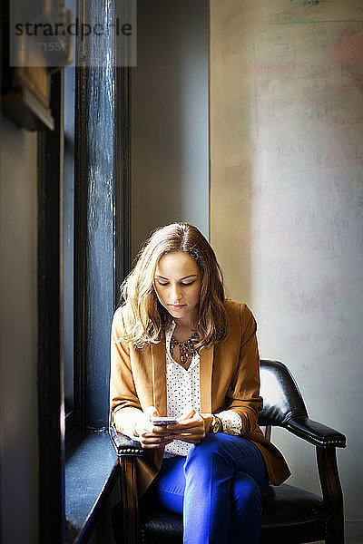 Frau benutzt Smartphone  während sie im Büro am Fenster sitzt