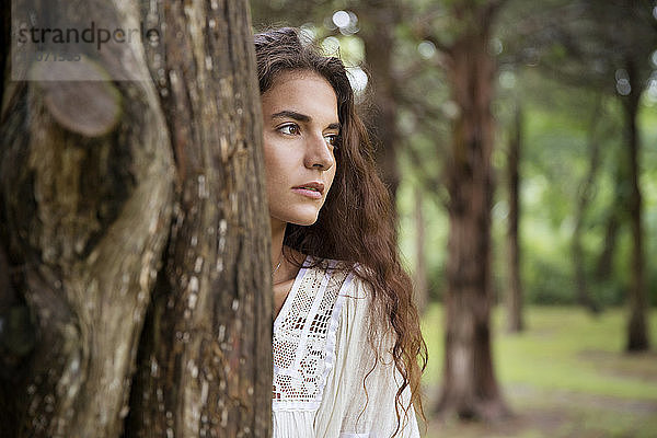 Frau schaut weg  während sie hinter einem Baumstamm im Wald steht