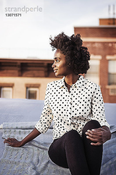 Lächelnde Frau sitzt auf Gebäudeterrasse gegen den Himmel
