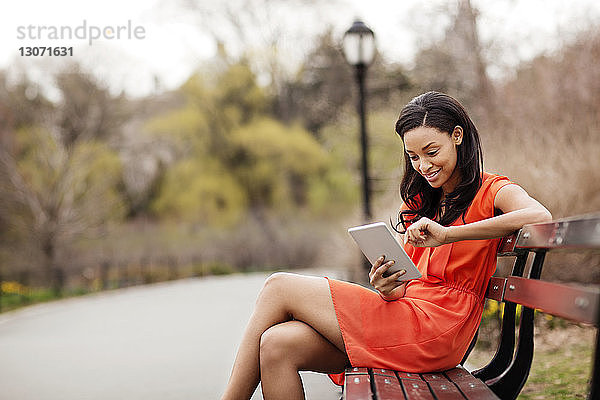 Glückliche Frau  die einen Tablet-Computer benutzt  während sie im Park auf einer Bank sitzt