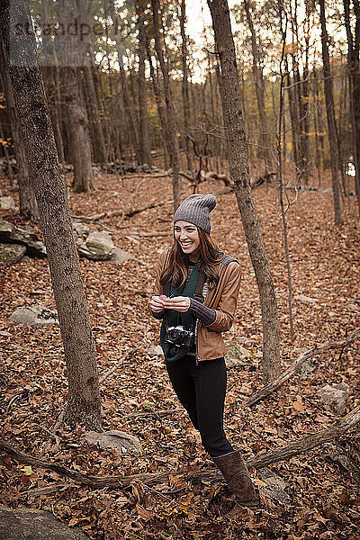 Lächelnde Frau mit Kamera steht im Winter inmitten von Bäumen im Wald