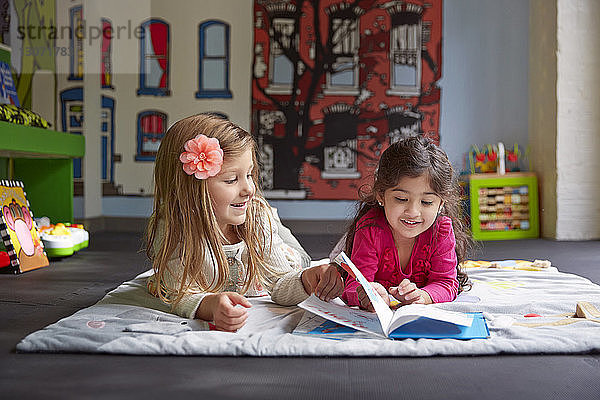 Lächelnde Freunde lesen ein Buch  während sie bei der Kinderbetreuung auf einer Decke liegen