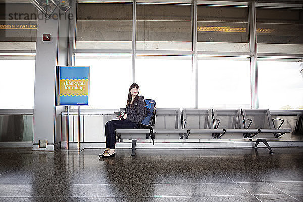 Frau mit Tablet-Computer schaut weg  während sie auf Sitzen im Warteraum des Bahnhofs sitzt