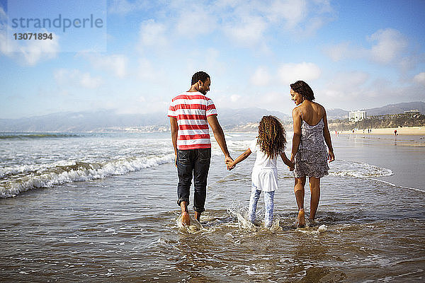 Rückansicht einer Familie mit Händchenhalten beim Strandspaziergang am Ufer