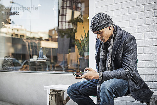 Mann benutzt Mobiltelefon  während er auf einer Bank an der Wand sitzt