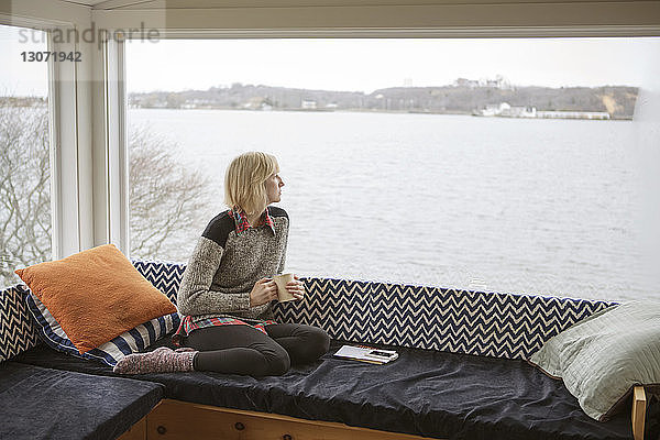 Nachdenkliche Frau trinkt im Winter Kaffee  während sie durch das Fenster auf den See schaut