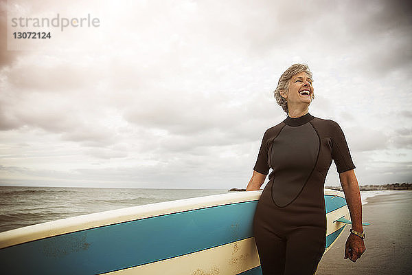 Glückliche Frau trägt ein Paddelbrett  während sie am Strand gegen den Himmel steht