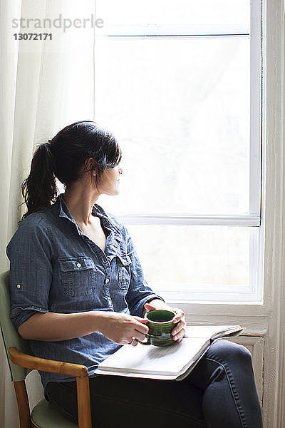 Frau mit Tasse und Buch  die wegschaut  während sie zu Hause auf einem Stuhl am Fenster sitzt