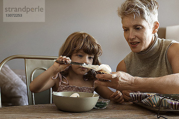Mädchen cremt Muffin ein  das von der Großmutter gehalten wird  während sie zu Hause sitzt