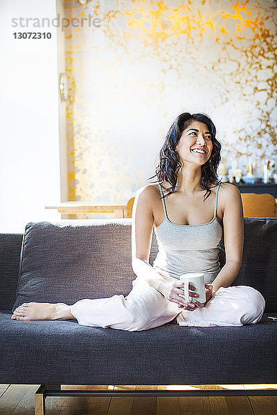 Glückliche Frau hält Kaffeetasse in der Hand und schaut weg  während sie sich auf dem Sofa entspannt