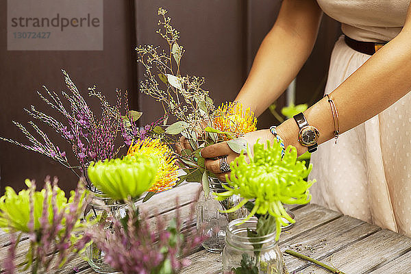 Beschnittenes Bild einer Frau  die Blumen in Gläsern auf einem Holztisch arrangiert