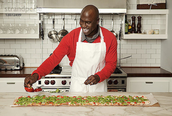 Fröhlicher Mann garniert Pizza auf Kücheninsel zu Hause