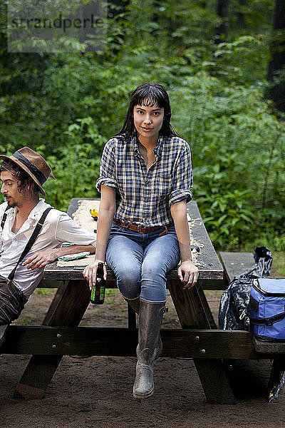 Porträt einer Frau  die eine Bierflasche hält  während sie mit einem Freund auf einem Picknicktisch im Wald sitzt