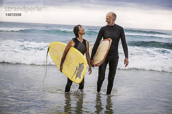 Glückliches Paar mit Surfbrettern im Gespräch beim Strandspaziergang am Ufer