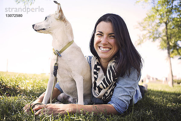 Porträt einer lächelnden Frau  die mit einem Hund auf einem Grasfeld im Park liegt