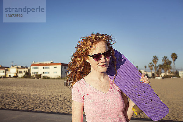 Frau hält Skateboard in der Hand  während sie am Strand vor strahlend blauem Himmel steht