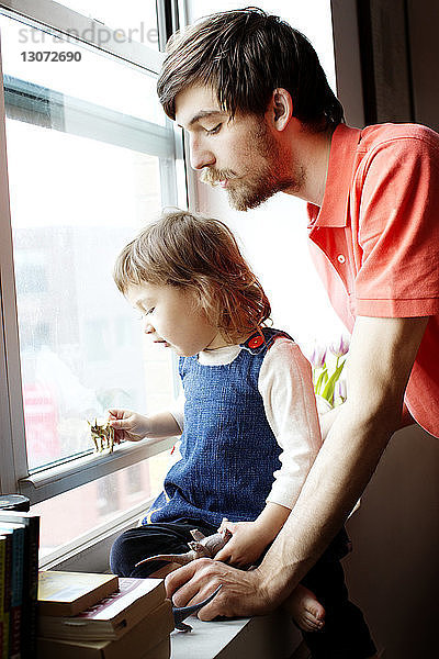 Vater sieht Mädchen an  das auf dem Fensterbrett sitzend mit Spielzeug spielt