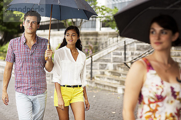 Freunde tragen Regenschirm beim Spaziergang auf einem Fußweg in der Stadt