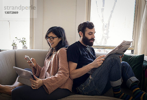 Frau benutzt Mobiltelefon  während der Ehemann auf dem Sofa Zeitung liest