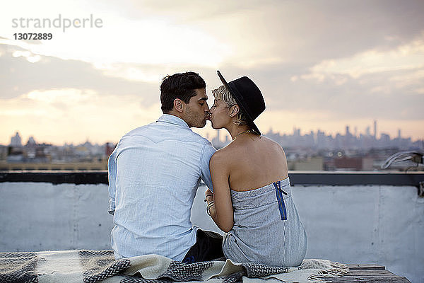 Rückansicht eines Paares  das sich küsst  während es bei Sonnenuntergang auf der Gebäudeterrasse sitzt
