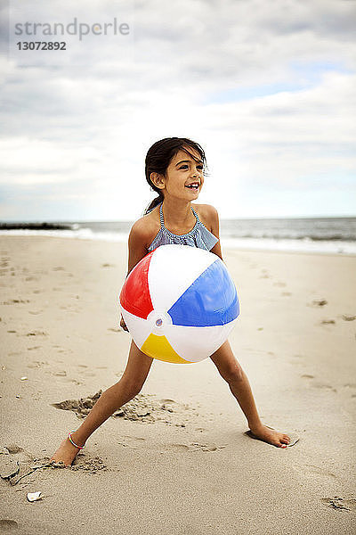Glückliches Mädchen spielt mit dem Ball  während es am Strand vor bewölktem Himmel steht